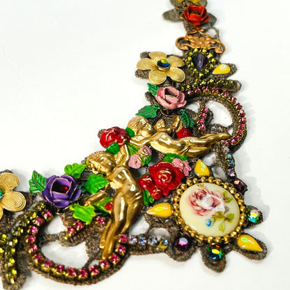 Detalle de collar artesanal con querubines, flores multicolor y cristales sobre encaje.